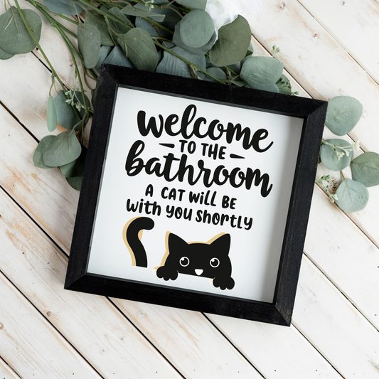 Welcome to the bathroom, a cat will be with you shortly framed sign with a black frame on white wood planks with eucalyptus