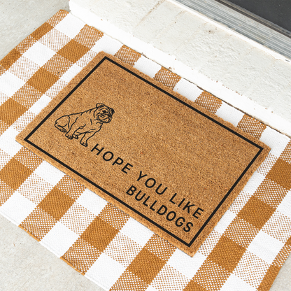 Hope You Like Bulldogs coir doormat with a drawing of a sitting bulldog at an angle on a tan and white check rug