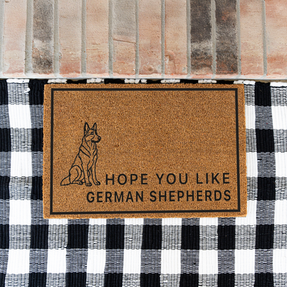 Hope You Like German Shepherds coir doormat with a drawing of a sitting German shepherd on a black and white buffalo check rug at a brick step