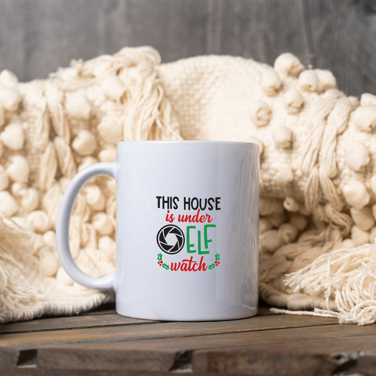 This House Is Under Elf Watch white ceramic Christmas coffee mug next to a cream knit blanket