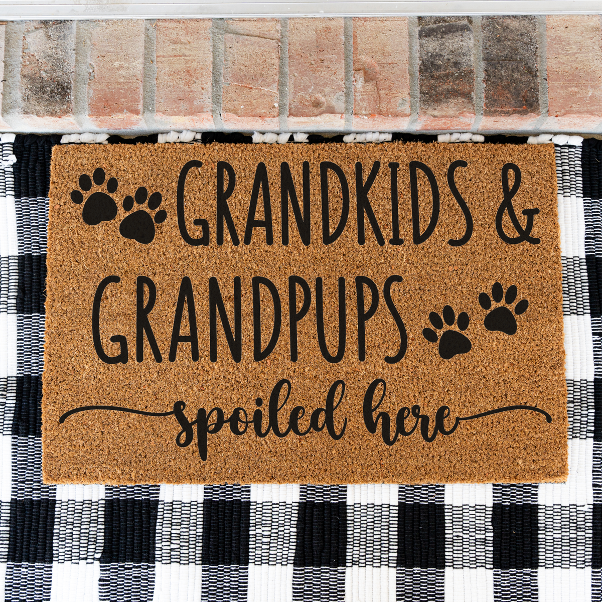 Grandkids & Grandpups Spoiled Here coir doormat with paw prints on a black and white buffalo check rug, close view