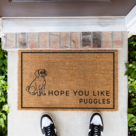Hope You Like Puggles coir doormat with a drawing of a sitting Puggle in front of a brick step, top-down view with sneakers