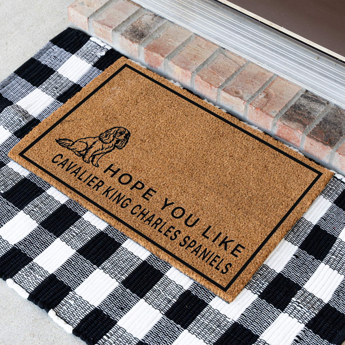 Hope You Like Cavalier King Charles Spaniels coir doormat with a drawing of a sitting Cavalier King Charles spaniel at an angle on a black and white buffalo check rug