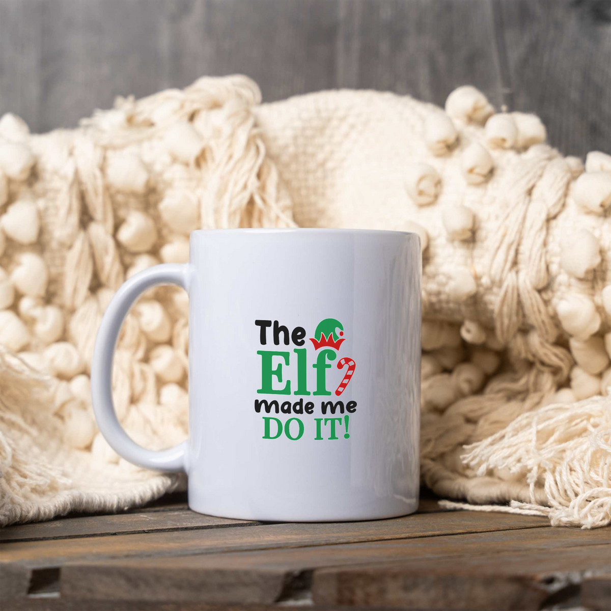 The Elf Made Me Do It white ceramic Christmas coffee mug next to a cream knit blanket