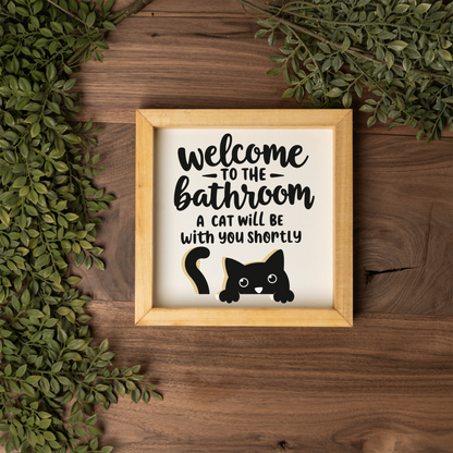 Welcome to the bathroom, a cat will be with you shortly framed sign with a natural wood frame on a dark wood table with greenery