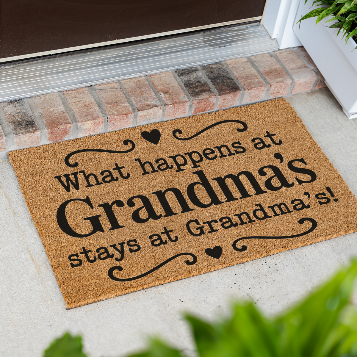 What Happens at Grandma's Stays at Grandma's coir doormat with hearts at an angle on a front porch by a brick step