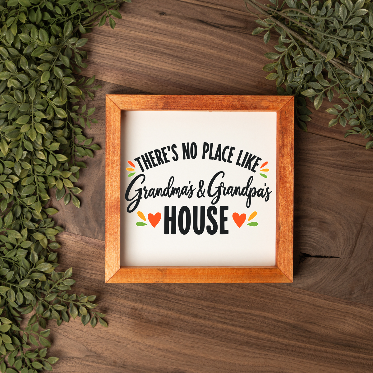 There's No Place Like Grandma's & Grandpa's House square framed sign with a stained wood frame on a dark wood table with greenery