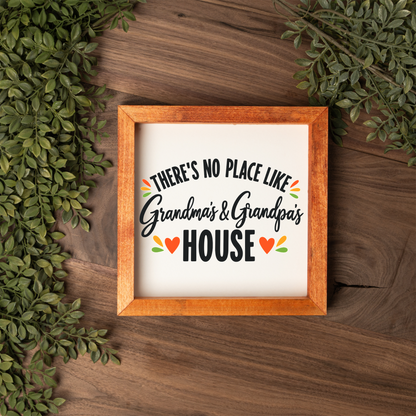 There's No Place Like Grandma's & Grandpa's House square framed sign with a stained wood frame on a dark wood table with greenery