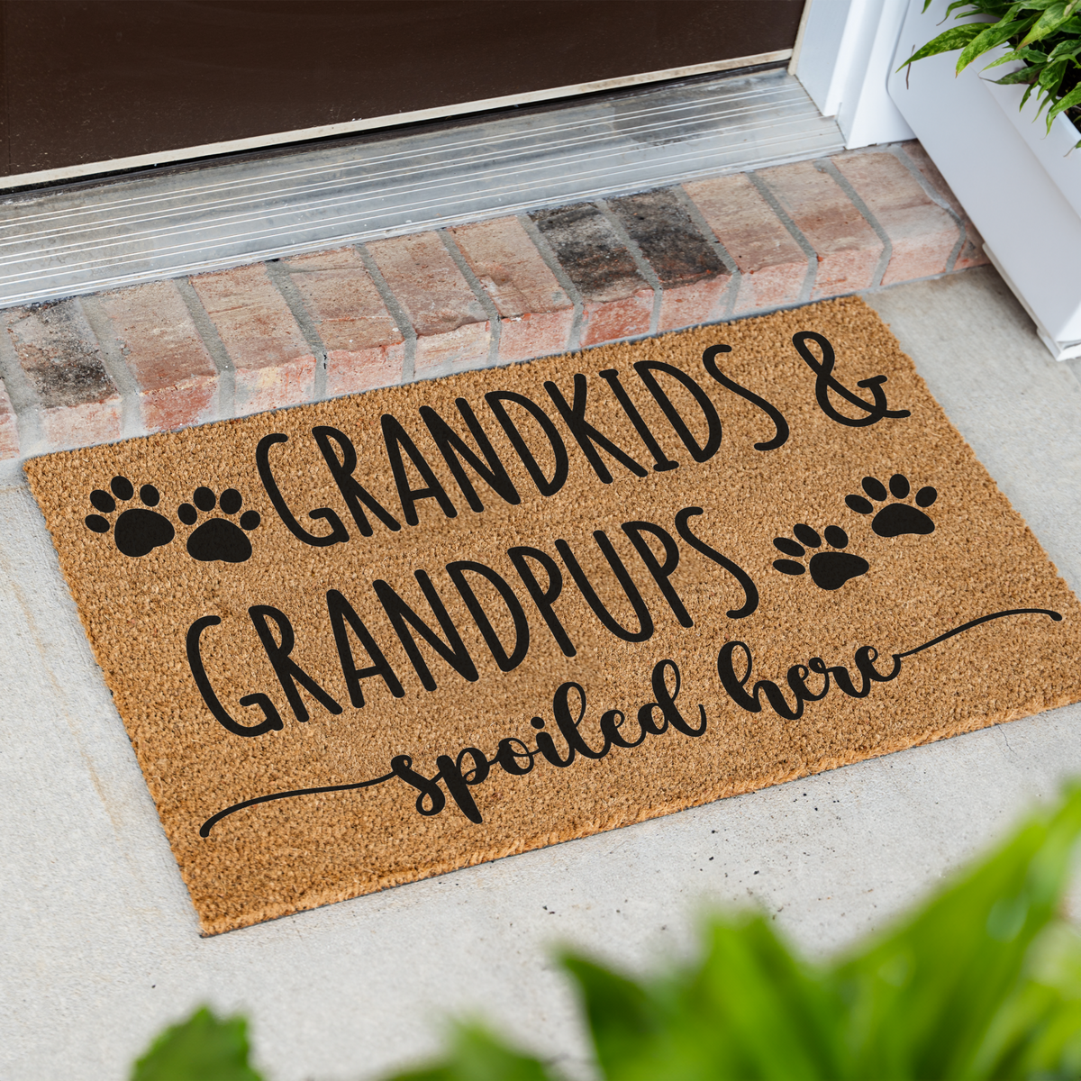 Grandkids & Grandpups Spoiled Here coir doormat with paw prints at an angle on a front porch by a brick step