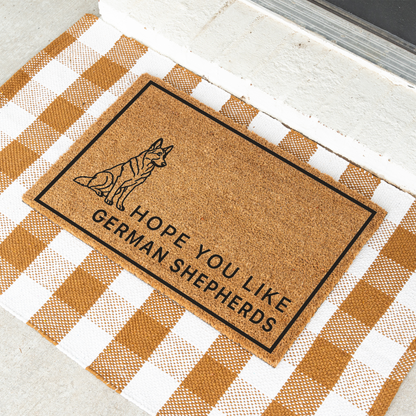 Hope You Like German Shepherds coir doormat with a drawing of a sitting German shepherd at an angle on a tan and white check rug