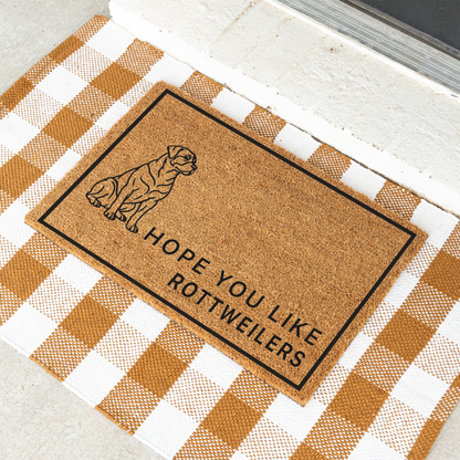 Hope You Like Rottweilers coir doormat with a drawing of a sitting Rottweiler at an angle on a tan and white check rug