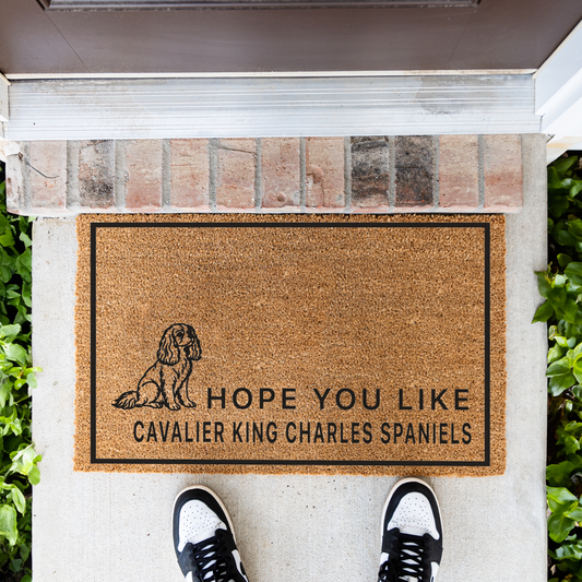 Hope You Like Cavalier King Charles Spaniels coir doormat with a drawing of a sitting Cavalier King Charles spaniel in front of a brick step, top-down view with sneakers