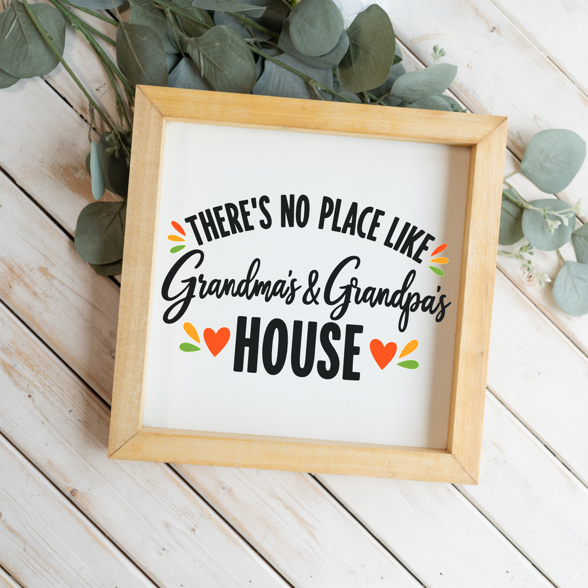 There's No Place Like Grandma's & Grandpa's House square framed sign with a natural wood frame on white wood planks with eucalyptus