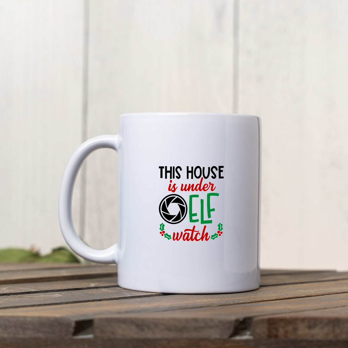 This House Is Under Elf Watch white ceramic Christmas coffee mug on a wooden crate against a white wood wall