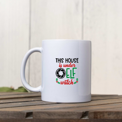 This House Is Under Elf Watch white ceramic Christmas coffee mug on a wooden crate against a white wood wall