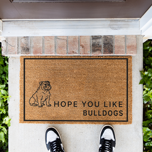 Hope You Like Bulldogs coir doormat with a drawing of a sitting bulldog in front of a brick step, top-down view with sneakers