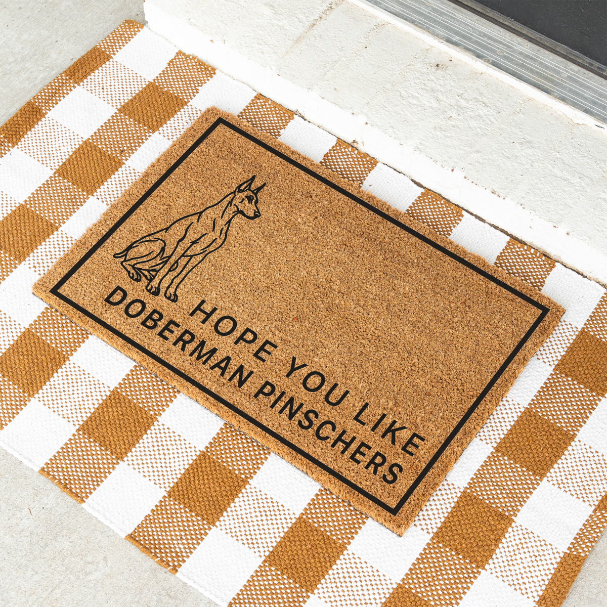 Hope You Like Doberman Pinschers coir doormat with a drawing of a sitting Doberman pinscher at an angle on a tan and white check rug