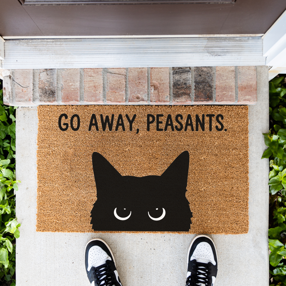 Go Away, Peasants black cat coir doormat in front of a brick step, top-down view