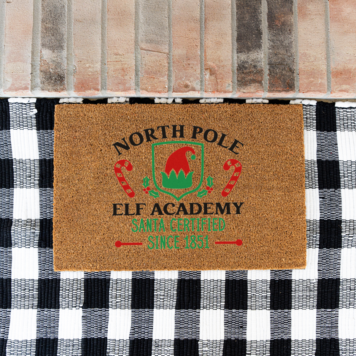 North Pole Elf Academy coir doormat on a black and white buffalo check rug at a brick step