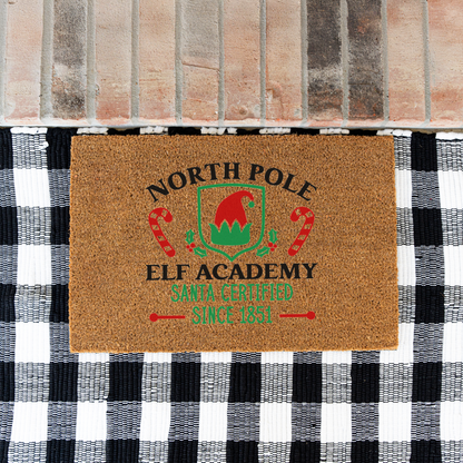 North Pole Elf Academy coir doormat on a black and white buffalo check rug at a brick step