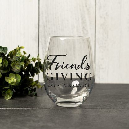 Friendsgiving stemless wine glass, empty, on a gray table by a white wood wall with greenery