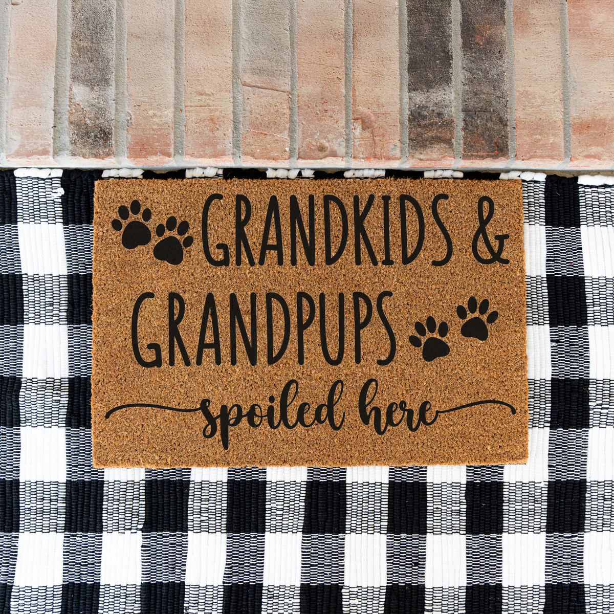 Grandkids & Grandpups Spoiled Here coir doormat with paw prints on a black and white buffalo check rug at a brick step