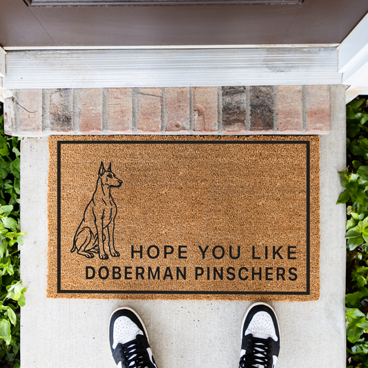 Hope You Like Doberman Pinschers coir doormat with a drawing of a sitting Doberman pinscher in front of a brick step, top-down view with sneakers