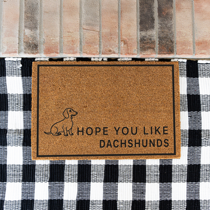Hope You Like Dachshunds coir doormat with a drawing of a sitting dachshund on a black and white buffalo check rug at a brick step