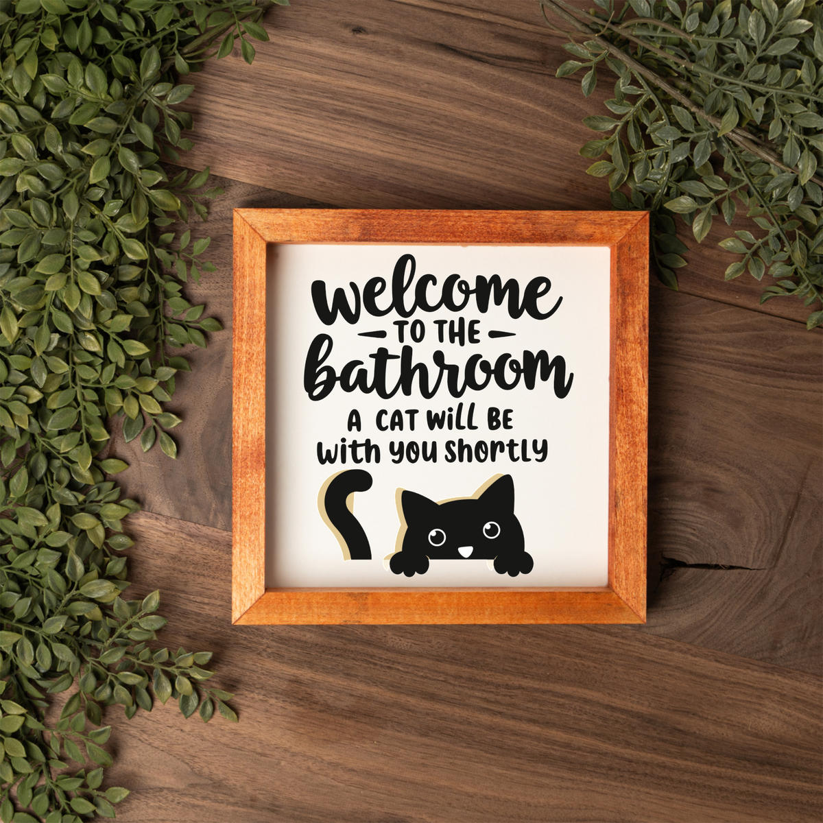 Welcome to the bathroom, a cat will be with you shortly framed sign with a stained wood frame on a dark wood table with greenery