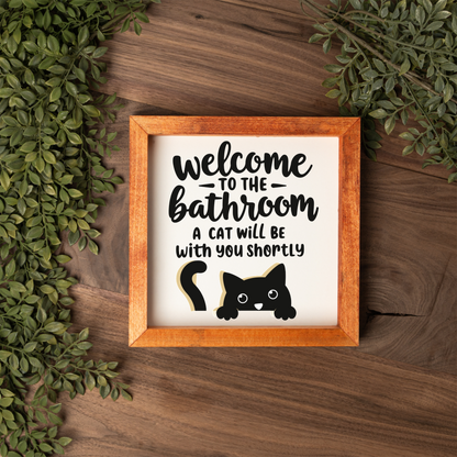Welcome to the bathroom, a cat will be with you shortly framed sign with a stained wood frame on a dark wood table with greenery