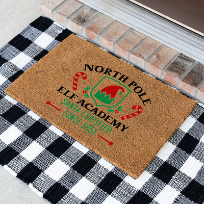 North Pole Elf Academy coir doormat at an angle on a black and white buffalo check rug
