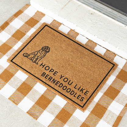 Hope You Like Bernedoodles coir doormat with a drawing of a sitting Bernedoodle at an angle on a tan and white check rug