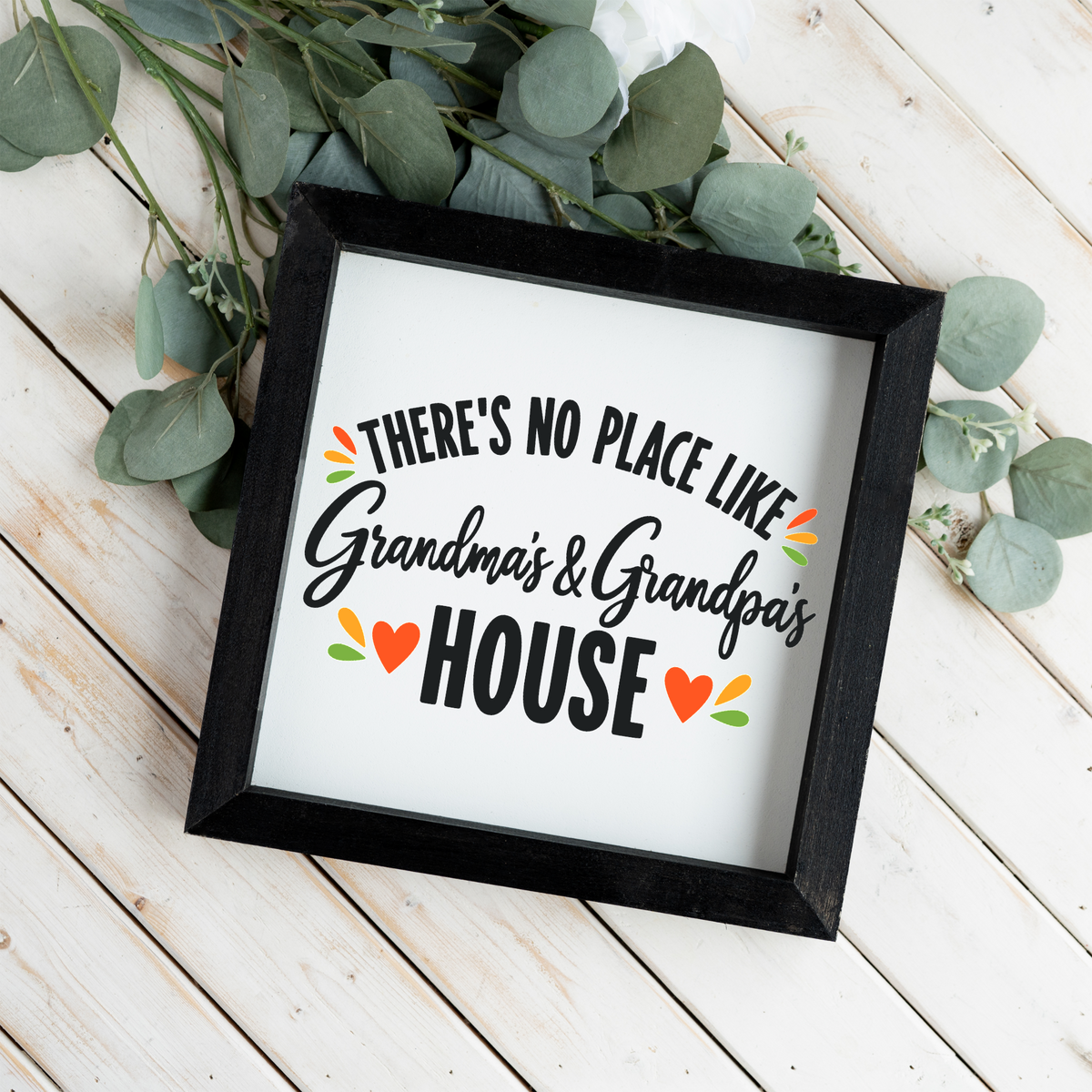 There's No Place Like Grandma's & Grandpa's House square framed sign with a black frame on white wood planks with eucalyptus