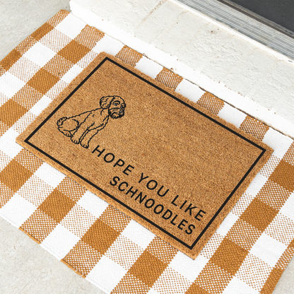 Hope You Like Schnoodles coir doormat with a drawing of a sitting Schnoodle at an angle on a tan and white check rug