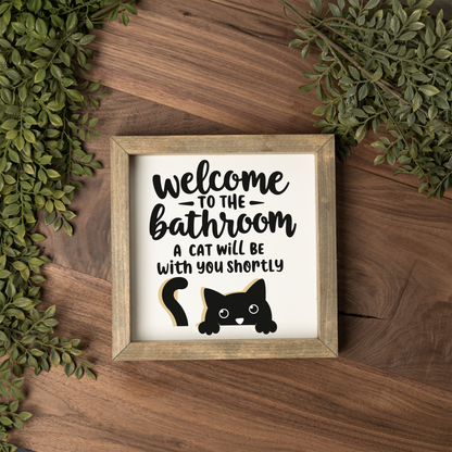 Welcome to the bathroom, a cat will be with you shortly framed sign with a weathered wood frame on a dark wood table with greenery