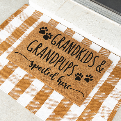 Grandkids & Grandpups Spoiled Here coir doormat with paw prints on a tan and white check rug