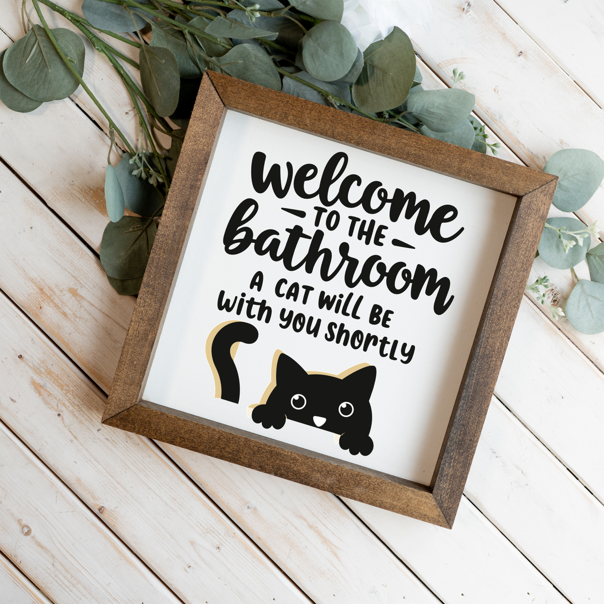 Welcome to the bathroom, a cat will be with you shortly framed sign with a walnut frame on white wood planks with eucalyptus