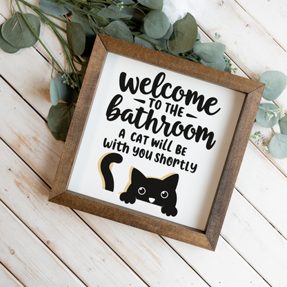 Welcome to the bathroom, a cat will be with you shortly framed sign with a walnut frame on white wood planks with eucalyptus