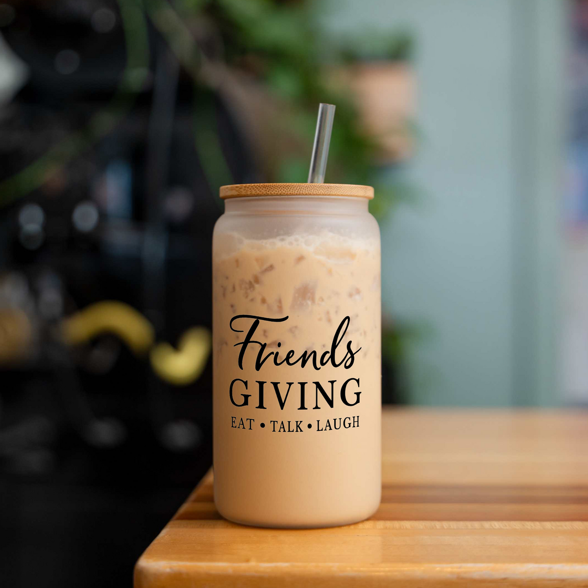 Friendsgiving 16 oz frosted glass can with a bamboo lid and straw, filled with iced coffee, on a wood table