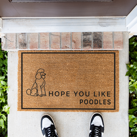 Hope You Like Poodles coir doormat with a drawing of a sitting poodle in front of a brick step, top-down view with sneakers