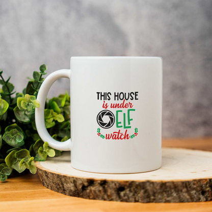 This House Is Under Elf Watch white ceramic Christmas coffee mug on a wood slice with greenery