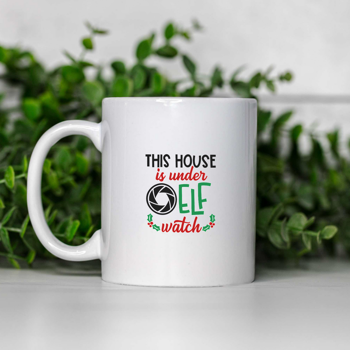 This House Is Under Elf Watch white ceramic Christmas coffee mug on a white shelf with greenery