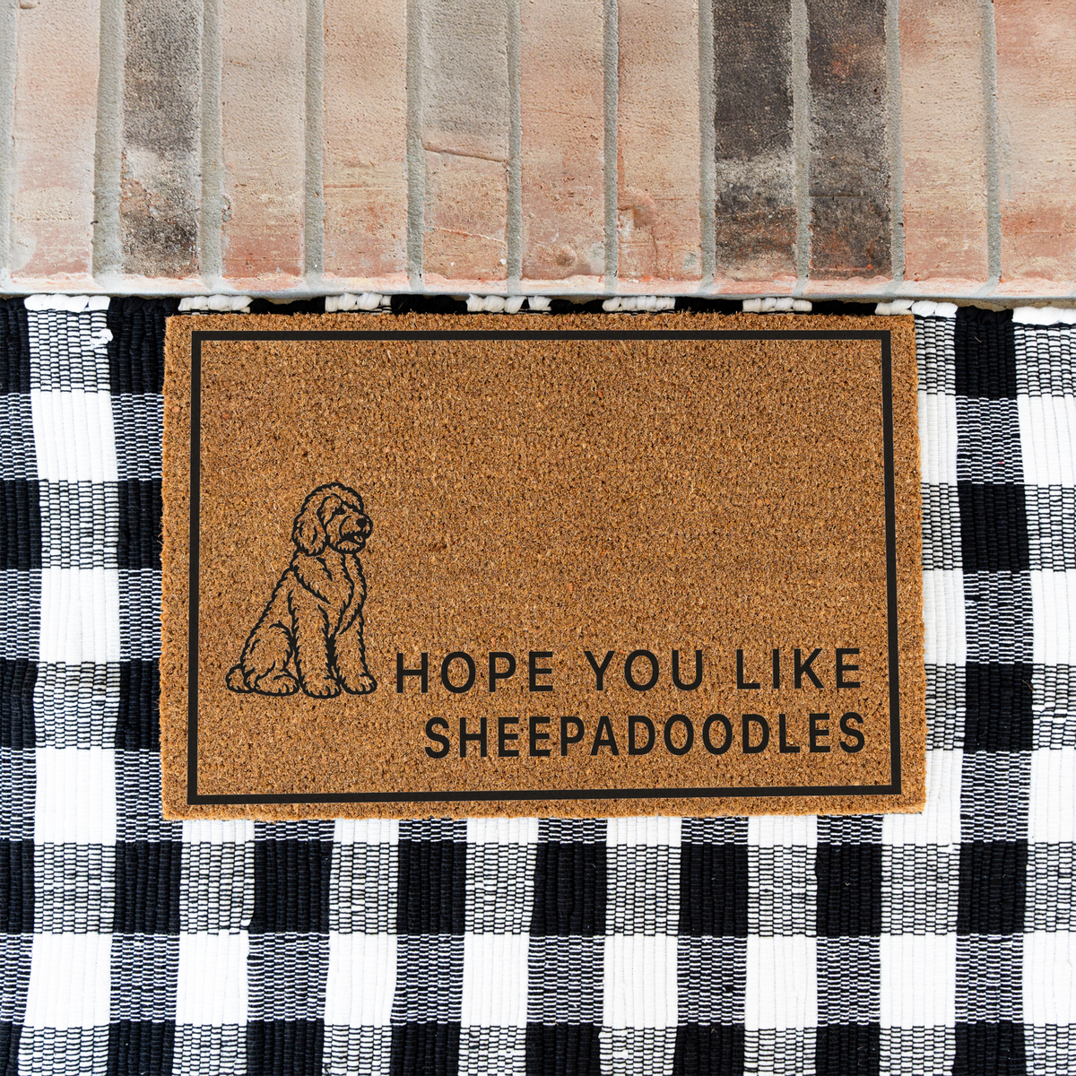 Hope You Like Sheepadoodles coir doormat with a drawing of a sitting Sheepadoodle on a black and white buffalo check rug at a brick step