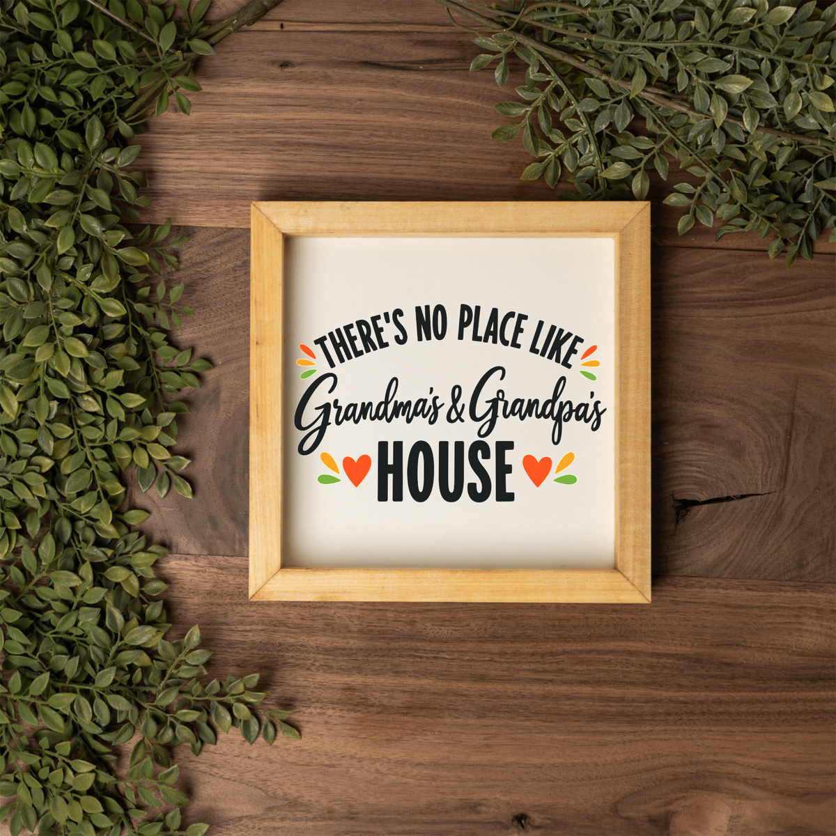 There's No Place Like Grandma's & Grandpa's House square framed sign with a natural wood frame on a dark wood table with greenery