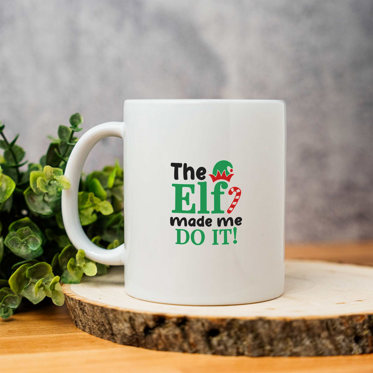 The Elf Made Me Do It white ceramic Christmas coffee mug on a wood slice with greenery