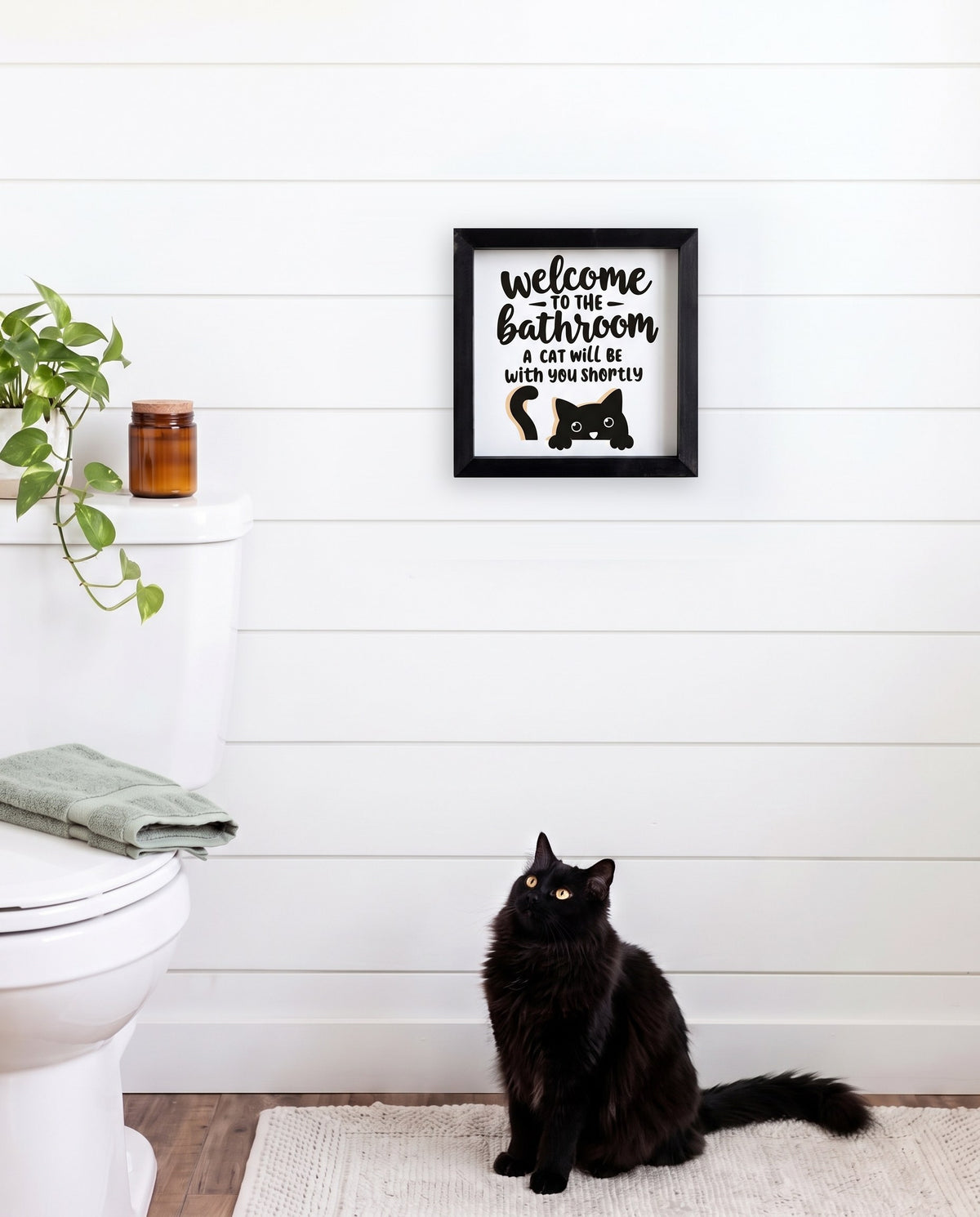 A Cat Will Be With You Shortly bathroom sign with a black frame, shown in 12x12, hanging on a white shiplap bathroom wall with a black cat below