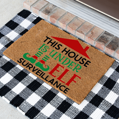 This House Is Under Elf Surveillance coir doormat at an angle on a black and white buffalo check rug