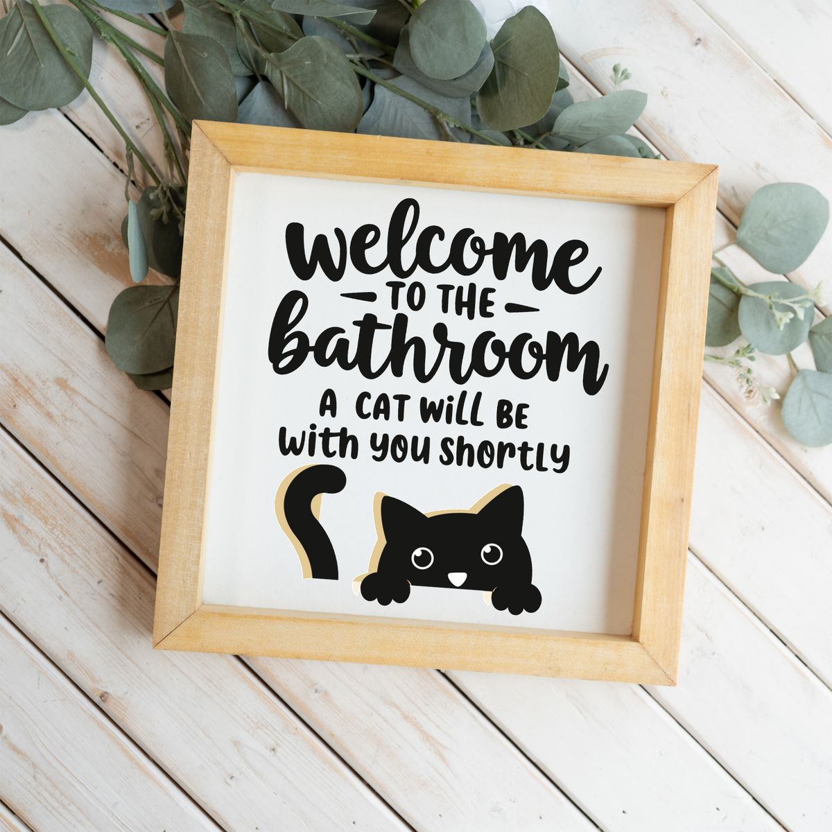Welcome to the bathroom, a cat will be with you shortly framed sign with a natural wood frame on white wood planks with eucalyptus
