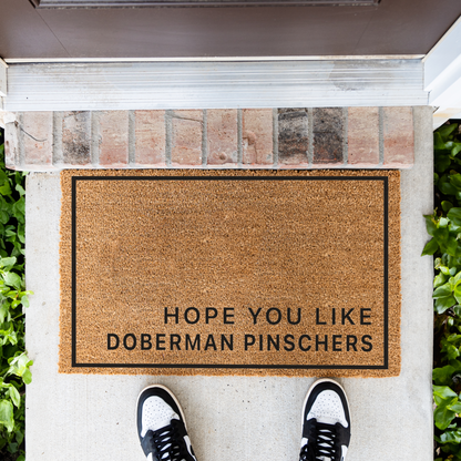 Hope You Like Doberman Pinschers minimalist coir doormat with a thin black border on a front porch, top-down view