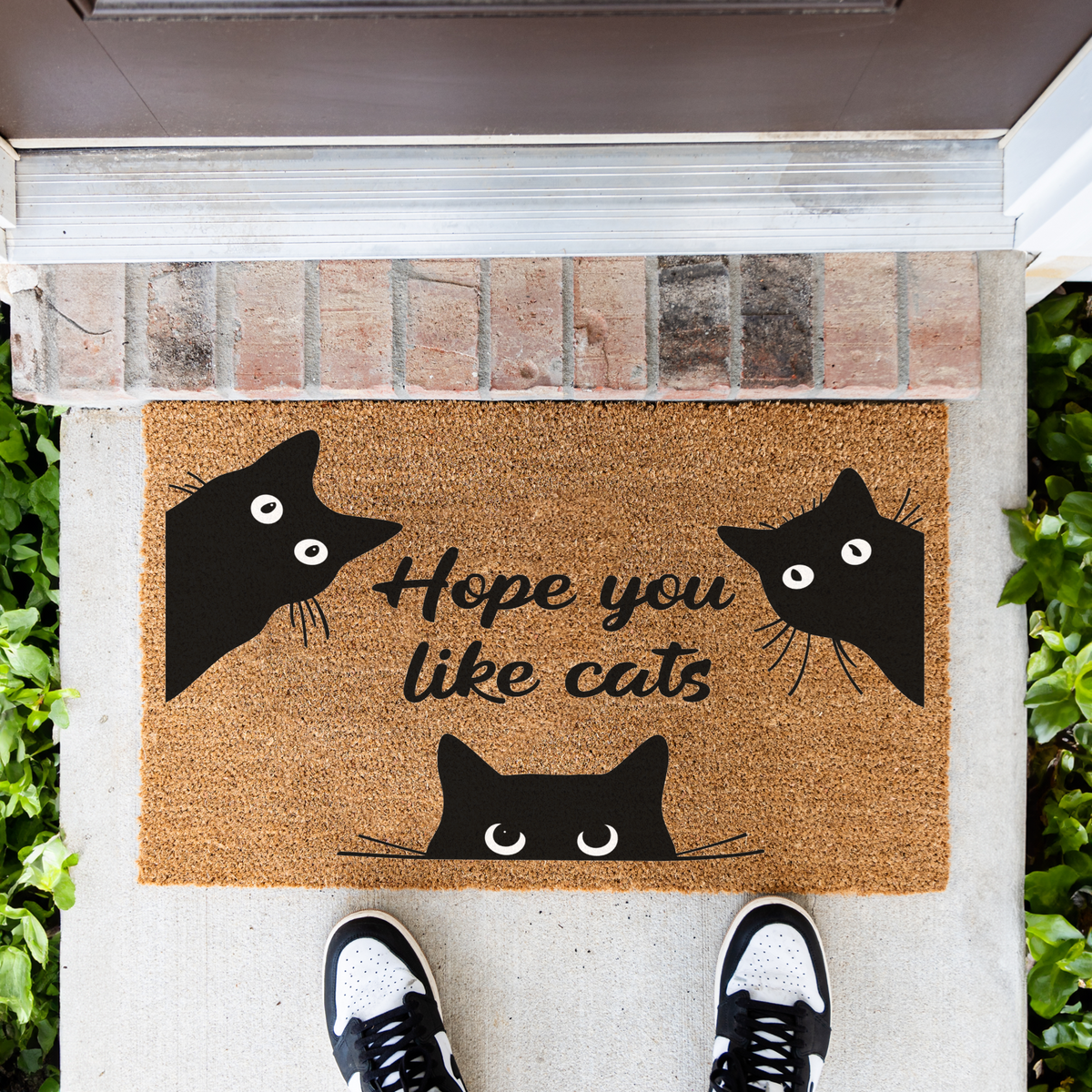 Hope You Like Cats coir doormat with three peeking black cats in front of a brick step, top-down view with sneakers